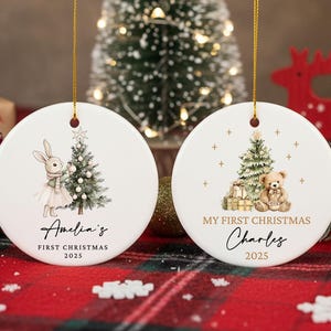 May include: Two white ceramic Christmas ornaments with gold hanging strings. One features a bunny with a Christmas tree, the text "Amelia's FIRST CHRISTMAS 2025". The other has a teddy bear, tree, gifts, and the text "MY FIRST CHRISTMAS Charles 2025".