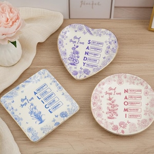 Puede incluir: Tres platos decorativos de cerámica con diseños florales y bordes dorados. Uno tiene forma de corazón, otro cuadrado y otro redondo. Cada plato presenta un nombre y palabras asociadas. Una peonía rosa y un jarrón están en el fondo.