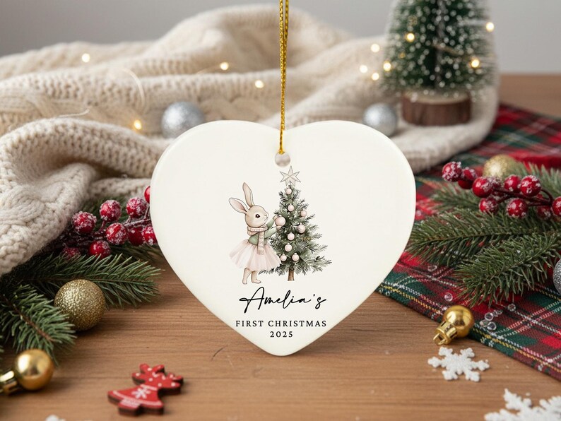 May include: Heart-shaped ceramic Christmas ornament with a bunny decorating a Christmas tree. The ornament has a gold string for hanging and features the text "Amelia's First Christmas 2025". Festive holiday decor.
