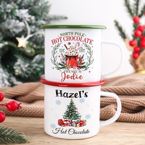 May include: Two white enamel mugs stacked on top of each other. The top mug has a green rim and reads "North Pole Hot Chocolate Premium Jodie." The bottom mug has a red rim and reads "Hazel's Hot Chocolate" with a Christmas tree design.