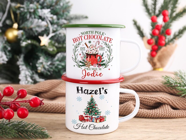 May include: Two white enamel mugs stacked on top of each other. The top mug has a green rim and reads "North Pole Hot Chocolate Premium Jodie." The bottom mug has a red rim and reads "Hazel's Hot Chocolate" with a Christmas tree design.