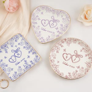 May include: Three personalized ceramic ring dishes in heart, square, and round shapes. Each dish features a floral design, two connected hearts with initials, and a date. The dishes have gold rims and are on a white surface.