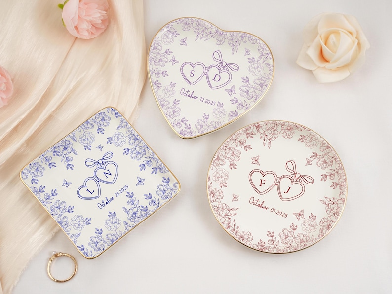 May include: Three personalized ceramic ring dishes in heart, square, and round shapes. Each dish features a floral design, two connected hearts with initials, and a date. The dishes have gold rims and are on a white surface.