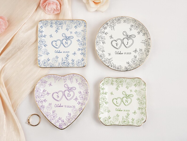 May include: Four decorative ring dishes in various shapes, each with a floral design and gold trim. The dishes feature intertwined heart shapes with initials and dates, including "October 28 2025" and "October 01 2025". A gold ring rests near the heart-shaped dish.