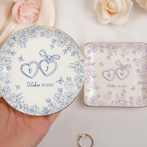 May include: Two decorative ceramic ring dishes. One round, one square. Both feature floral designs and heart-shaped elements with initials and dates. The round dish has blue floral patterns and the square dish has purple floral patterns.