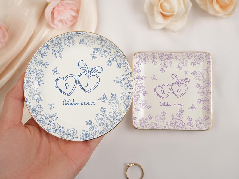 May include: Two decorative ceramic ring dishes. One round, one square. Both feature floral designs and heart-shaped elements with initials and dates. The round dish has blue floral patterns and the square dish has purple floral patterns.