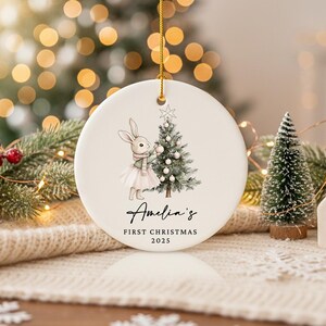May include: A round, white Christmas ornament with a gold string. The ornament features a watercolor illustration of a bunny wearing a scarf and tutu, decorating a Christmas tree. Text reads "Amelia's First Christmas 2025."
