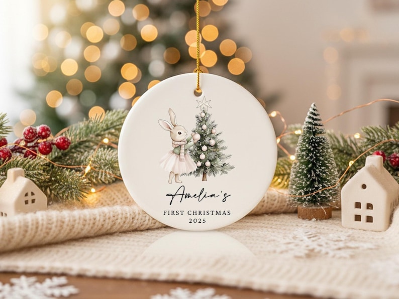 May include: A round, white Christmas ornament with a gold string. The ornament features a watercolor illustration of a bunny wearing a scarf and tutu, decorating a Christmas tree. Text reads "Amelia's First Christmas 2025."