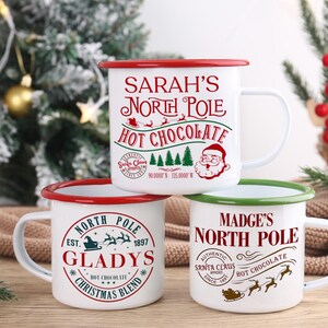 May include: Three white enamel mugs with red and green rims are stacked. Each mug has a different design with text related to the North Pole and hot chocolate. The mugs are on a wooden surface with a Christmas tree in the background.