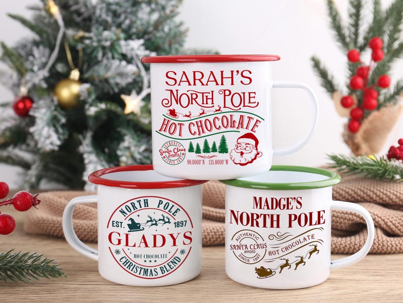May include: Three white enamel mugs with red and green rims are stacked. Each mug has a different design with text related to the North Pole and hot chocolate. The mugs are on a wooden surface with a Christmas tree in the background.