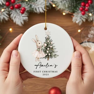 May include: A white ceramic Christmas ornament featuring a bunny decorating a Christmas tree. The ornament has the text "Amelia's First Christmas 2025" in script. A gold string is attached for hanging. The ornament is held by two hands.