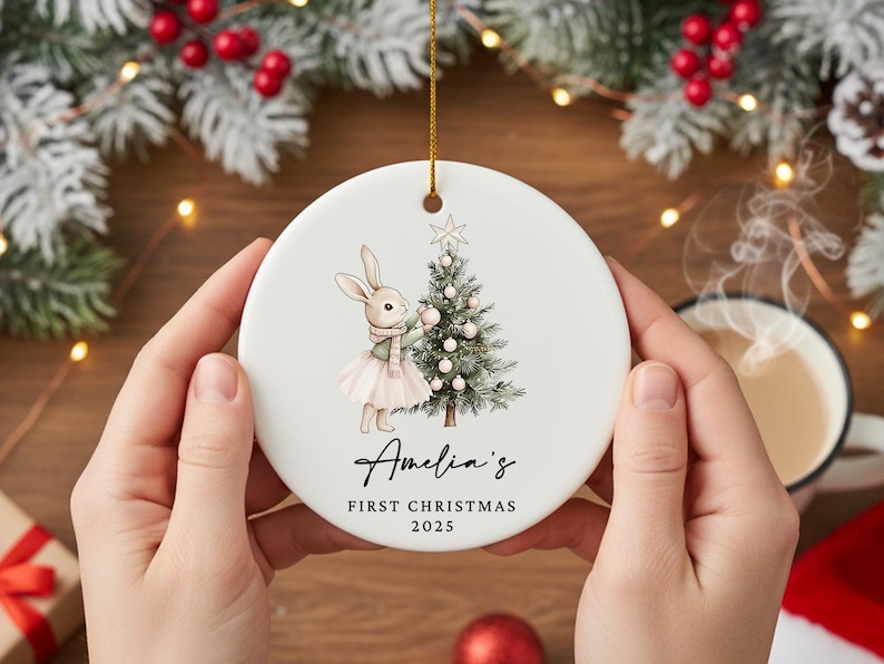 May include: A white ceramic Christmas ornament featuring a bunny decorating a Christmas tree. The ornament has the text "Amelia's First Christmas 2025" in script. A gold string is attached for hanging. The ornament is held by two hands.