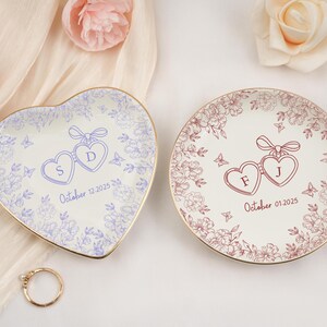 May include: Two decorative ring dishes, one heart-shaped and one round, with floral and heart designs. The heart-shaped dish has the letters "S" and "D" and the date "October 12, 2025". The round dish has the letters "F" and "J" and the date "October 01, 2025".