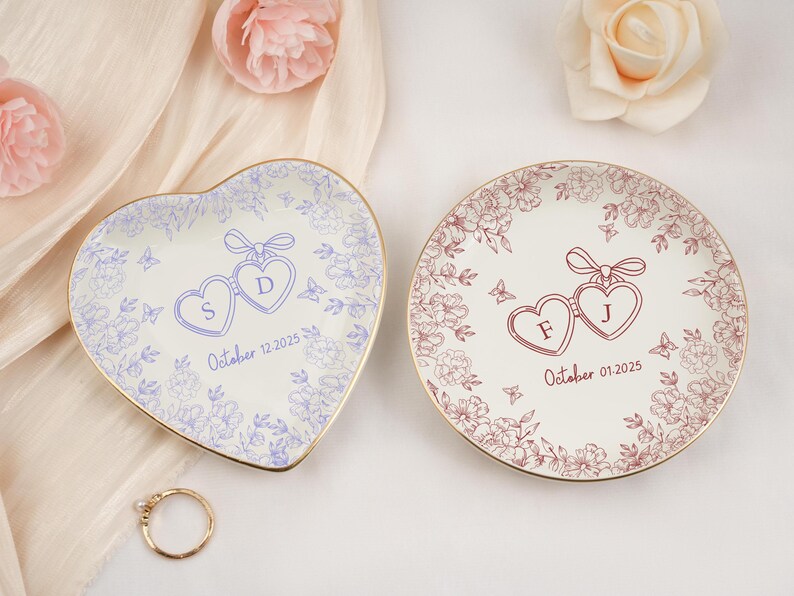 May include: Two decorative ring dishes, one heart-shaped and one round, with floral and heart designs. The heart-shaped dish has the letters "S" and "D" and the date "October 12, 2025". The round dish has the letters "F" and "J" and the date "October 01, 2025".