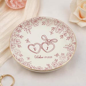 May include: A white ceramic ring dish with a gold rim, adorned with a floral pattern and two connected heart shapes. The hearts contain the letters "F" and "J", with the date "October 01.2025" below. Decorative butterflies are also present.