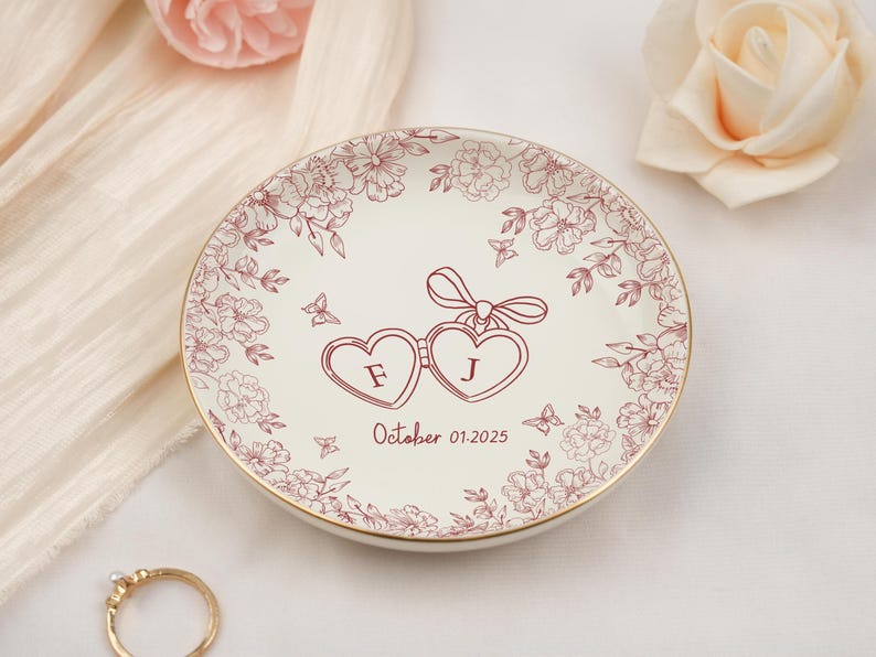May include: A white ceramic ring dish with a gold rim, adorned with a floral pattern and two connected heart shapes. The hearts contain the letters "F" and "J", with the date "October 01.2025" below. Decorative butterflies are also present.