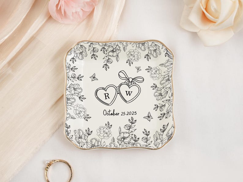 May include: Square, off-white ceramic ring dish with a gold trim. The dish features a floral design with two connected heart-shaped pendants, each with a letter inside. The date "October 25, 2025" is written below the hearts.