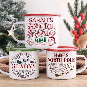 May include: Three white enamel mugs with red or green rims and handles are stacked. Each mug features a festive design with the words "North Pole" and "Hot Chocolate". One mug says "Sarah's", another says "Gladys", and the third says "Madge's".