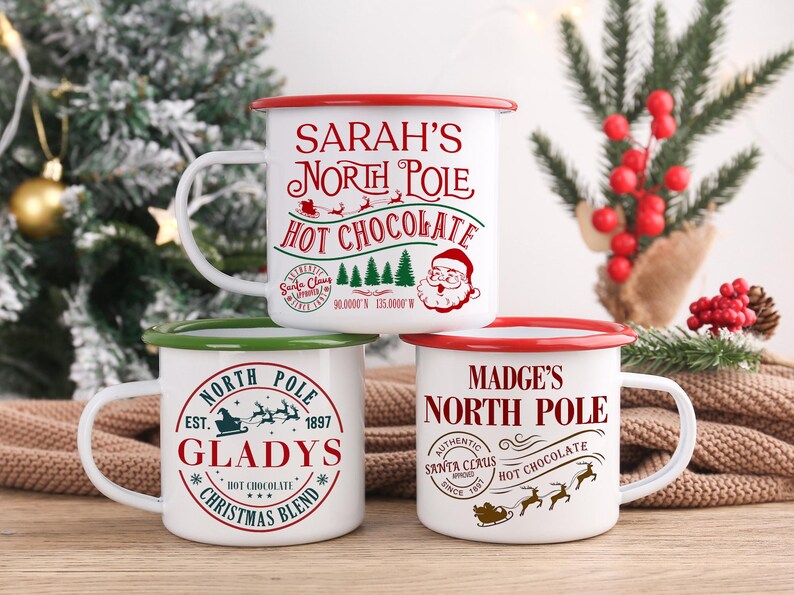 May include: Three white enamel mugs with red or green rims and handles are stacked. Each mug features a festive design with the words "North Pole" and "Hot Chocolate". One mug says "Sarah's", another says "Gladys", and the third says "Madge's".
