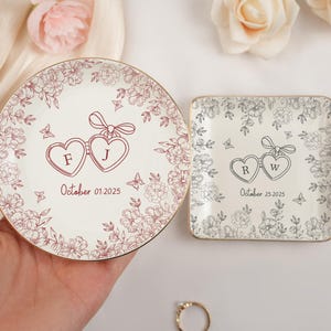 May include: Two personalized ceramic ring dishes. One round and one square, both with floral designs and gold rims. The round dish has the initials "F J" and the date "October 01.2025". The square dish has the initials "R W" and the date "October 25.2025".