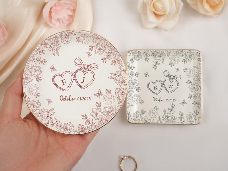 May include: Two personalized ceramic ring dishes. One round and one square, both with floral designs and gold rims. The round dish has the initials "F J" and the date "October 01.2025". The square dish has the initials "R W" and the date "October 25.2025".