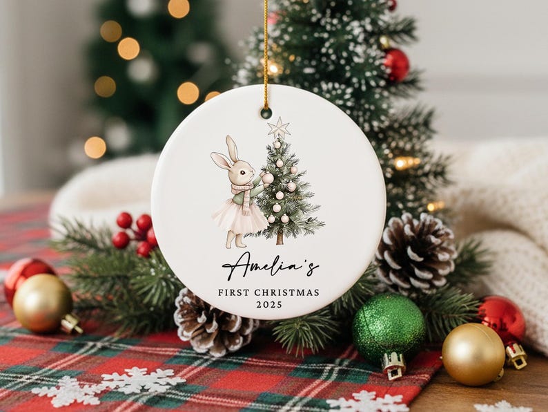 May include: A white ceramic Christmas ornament with a bunny and Christmas tree illustration. The ornament has the text "Amelia's First Christmas 2025". The ornament is hanging in front of a Christmas tree and decorations.
