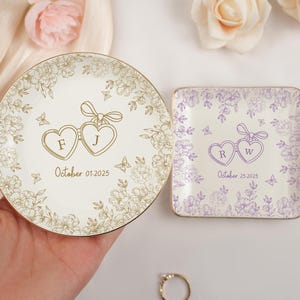 May include: Two personalized ring dishes. One round, one square. The round dish is cream-colored with gold floral accents, initials "F J", and the date "October 01, 2025". The square dish is lavender with initials "R W" and the date "October 25, 2025".