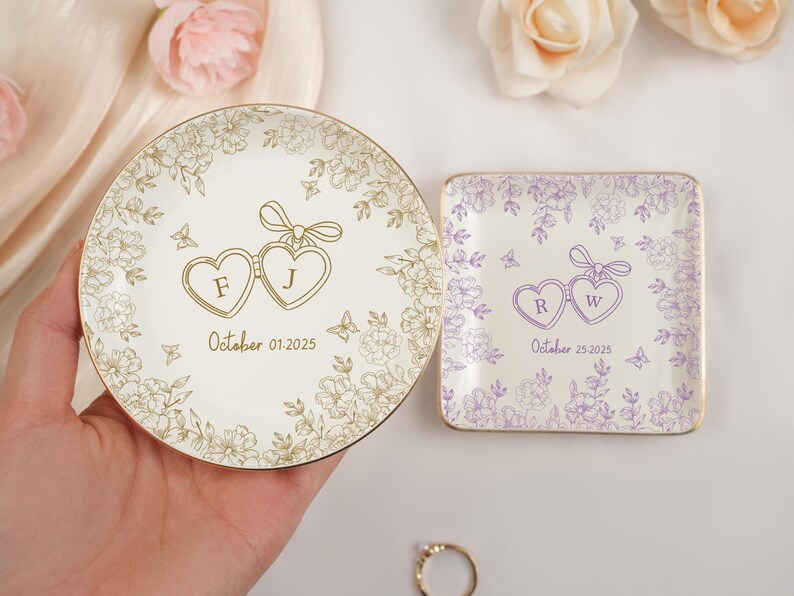 May include: Two personalized ring dishes. One round, one square. The round dish is cream-colored with gold floral accents, initials "F J", and the date "October 01, 2025". The square dish is lavender with initials "R W" and the date "October 25, 2025".