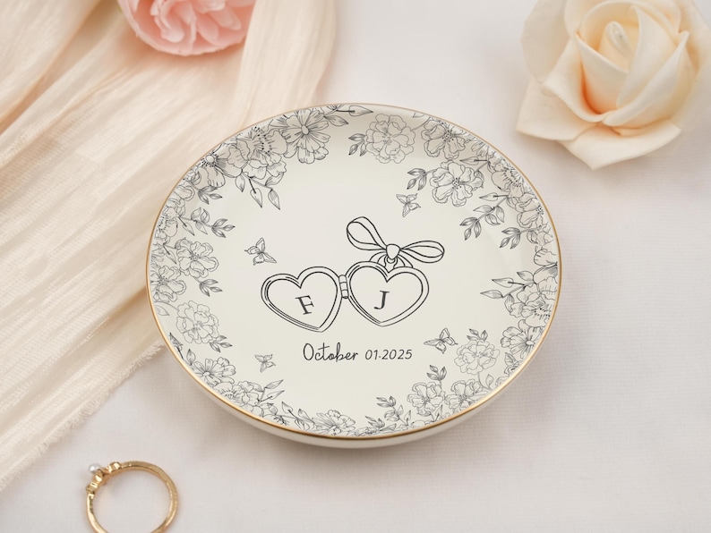 May include: A white ceramic ring dish with a gold rim, featuring a floral design and two heart-shaped frames with the letters "F" and "J". The date "October 01.2025" is printed below. Decorative butterflies are also included.