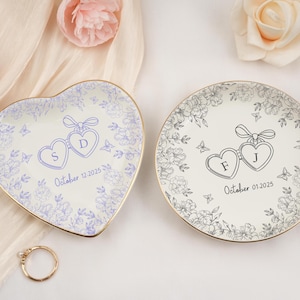 May include: Two personalized ring dishes, one heart-shaped and one round, with floral designs and gold rims. The heart-shaped dish has the initials "S" and "D" with the date "October 12, 2025". The round dish has the initials "F" and "J" with the date "October 01, 2025".