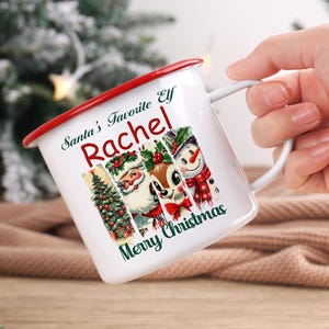 May include: White enamel mug with a red rim and handle, featuring a Christmas design. The mug has the text "Santa's Favorite Elf Rachel Merry Christmas" and images of a Christmas tree, Santa, a reindeer, and a snowman.