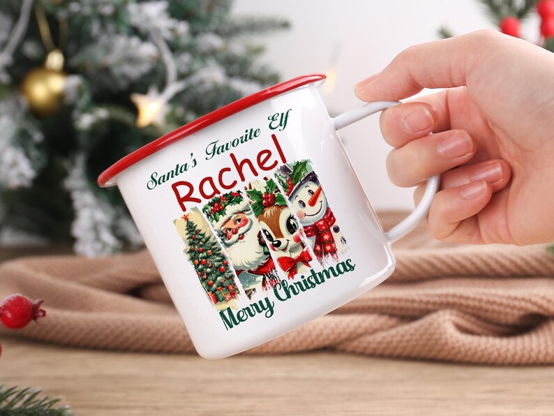 May include: White enamel mug with a red rim and handle, featuring a Christmas design. The mug has the text "Santa's Favorite Elf Rachel Merry Christmas" and images of a Christmas tree, Santa, a reindeer, and a snowman.