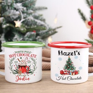 May include: Two white enamel mugs with Christmas-themed designs. One mug has a green rim and reads "North Pole Hot Chocolate" with a graphic of a hot chocolate. The other mug has a red rim and reads "Hazel's Hot Chocolate" with a Christmas tree graphic.