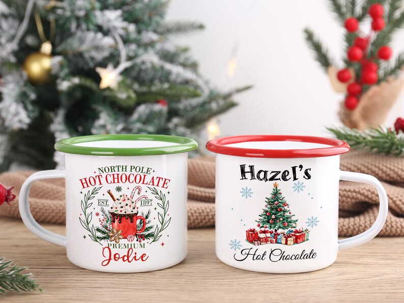 May include: Two white enamel mugs with Christmas-themed designs. One mug has a green rim and reads "North Pole Hot Chocolate" with a graphic of a hot chocolate. The other mug has a red rim and reads "Hazel's Hot Chocolate" with a Christmas tree graphic.