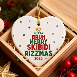 May include: A heart-shaped Christmas ornament with the words "67 NO CAP BRUH MERRY SKIBIDI RIZZMAS 2025" in red and green. The ornament is white with black snowflake accents and a gold string. It sits in a wooden box.