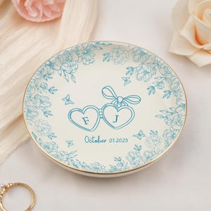 May include: A white ceramic ring dish with a gold rim, adorned with a teal floral design. The center features two heart-shaped glasses with the initials "F" and "J" and the date "October 01.2025".