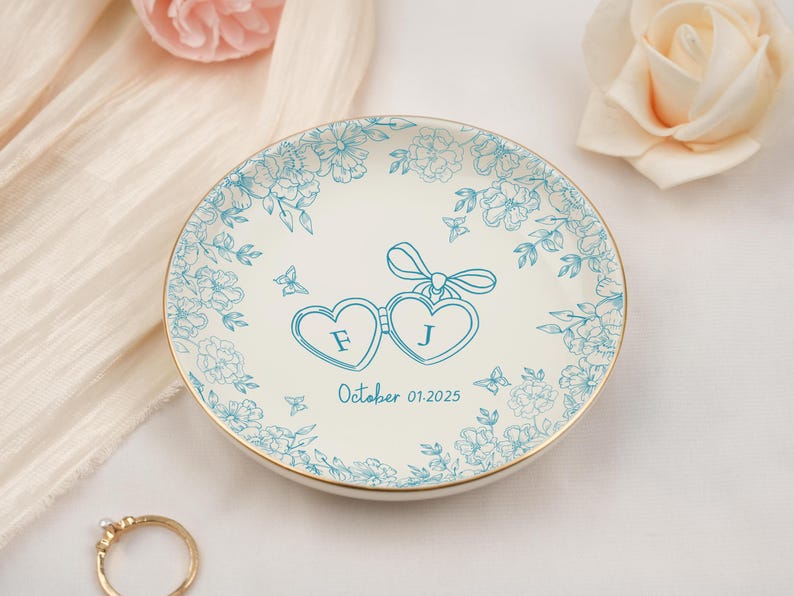 May include: A white ceramic ring dish with a gold rim, adorned with a teal floral design. The center features two heart-shaped glasses with the initials "F" and "J" and the date "October 01.2025".