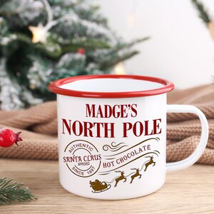 May include: White enamel mug with a red rim and handle. The mug features the text "MADGE'S NORTH POLE" in a festive font, along with a Santa Claus approved stamp and a graphic of Santa's sleigh and reindeer. The mug is designed for hot chocolate.