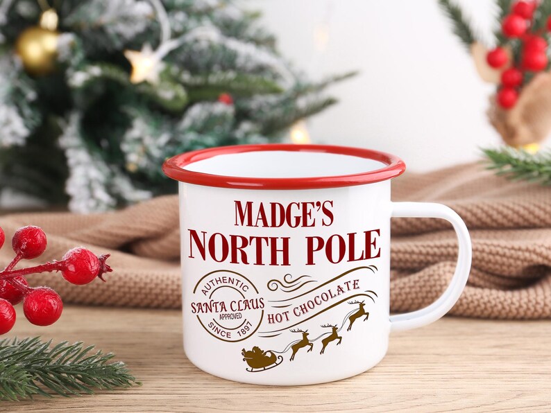May include: White enamel mug with a red rim and handle. The mug features the text "MADGE'S NORTH POLE" in a festive font, along with a Santa Claus approved stamp and a graphic of Santa's sleigh and reindeer. The mug is designed for hot chocolate.
