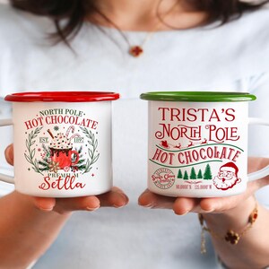 May include: Two white enamel mugs with red and green rims. One mug reads "North Pole Hot Chocolate" with a graphic of a hot chocolate drink. The other mug says "Trista's North Pole Hot Chocolate" with a Santa Claus graphic.