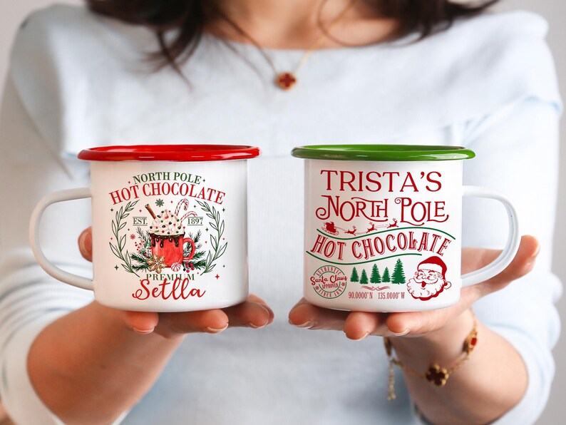 May include: Two white enamel mugs with red and green rims. One mug reads "North Pole Hot Chocolate" with a graphic of a hot chocolate drink. The other mug says "Trista's North Pole Hot Chocolate" with a Santa Claus graphic.