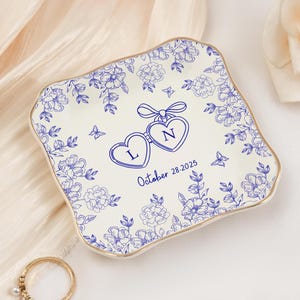 May include: A square, off-white ceramic dish with a blue floral design and gold trim. The dish features two connected heart-shaped pendants with the letters "L" and "N" and the date "October 28.2025".