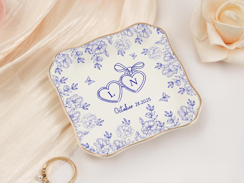 May include: A square, off-white ceramic dish with a blue floral design and gold trim. The dish features two connected heart-shaped pendants with the letters "L" and "N" and the date "October 28.2025".