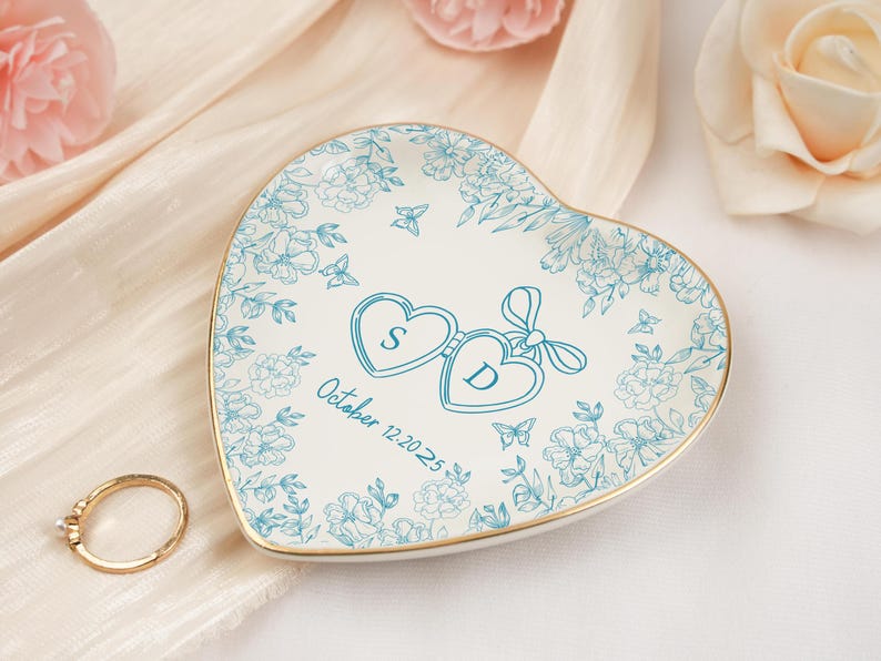 May include: Heart-shaped ring dish with a floral design in teal and gold trim. The dish features two hearts with the letters "S" and "D" and the date "October 12-20-2025". A gold ring rests beside the dish.