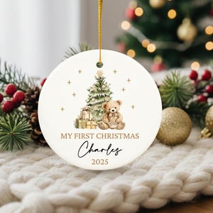 May include: A white Christmas ornament with a gold string, featuring a watercolor illustration of a Christmas tree, teddy bear, and gifts. Text reads "MY FIRST CHRISTMAS" and a name, with the year 2025.