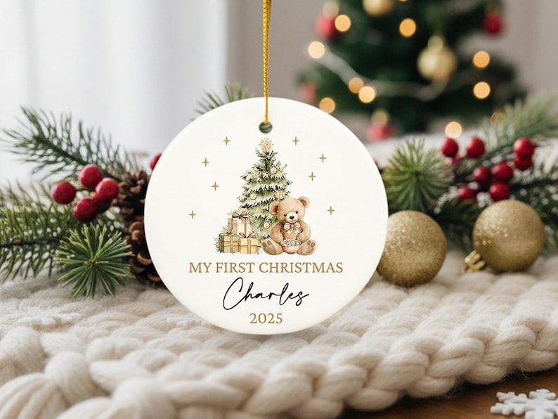 May include: A white Christmas ornament with a gold string, featuring a watercolor illustration of a Christmas tree, teddy bear, and gifts. Text reads "MY FIRST CHRISTMAS" and a name, with the year 2025.