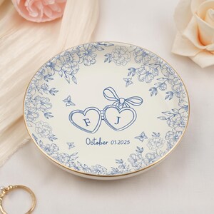 May include: A white ceramic ring dish with a gold rim, adorned with blue floral patterns and two heart-shaped frames with the letters "F" and "J". The date "October 01.2025" is also displayed. A charming wedding or engagement keepsake.