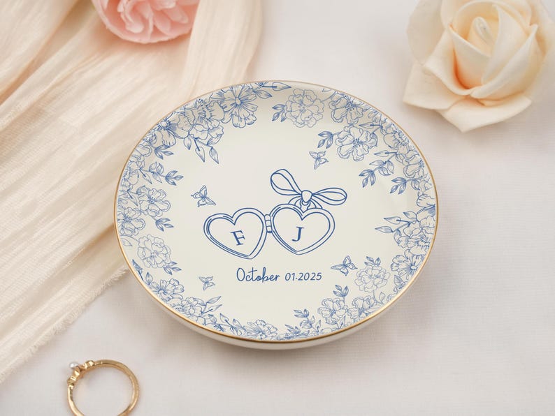 May include: A white ceramic ring dish with a gold rim, adorned with blue floral patterns and two heart-shaped frames with the letters "F" and "J". The date "October 01.2025" is also displayed. A charming wedding or engagement keepsake.