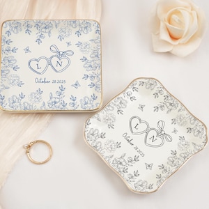 May include: Two decorative ceramic ring dishes with floral designs. One is square, the other is diamond-shaped. Both have gold rims, blue floral patterns, and heart-shaped initials with a date. A gold ring with a pearl is also present.
