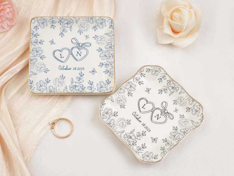 May include: Two decorative ceramic ring dishes with floral designs. One is square, the other is diamond-shaped. Both have gold rims, blue floral patterns, and heart-shaped initials with a date. A gold ring with a pearl is also present.